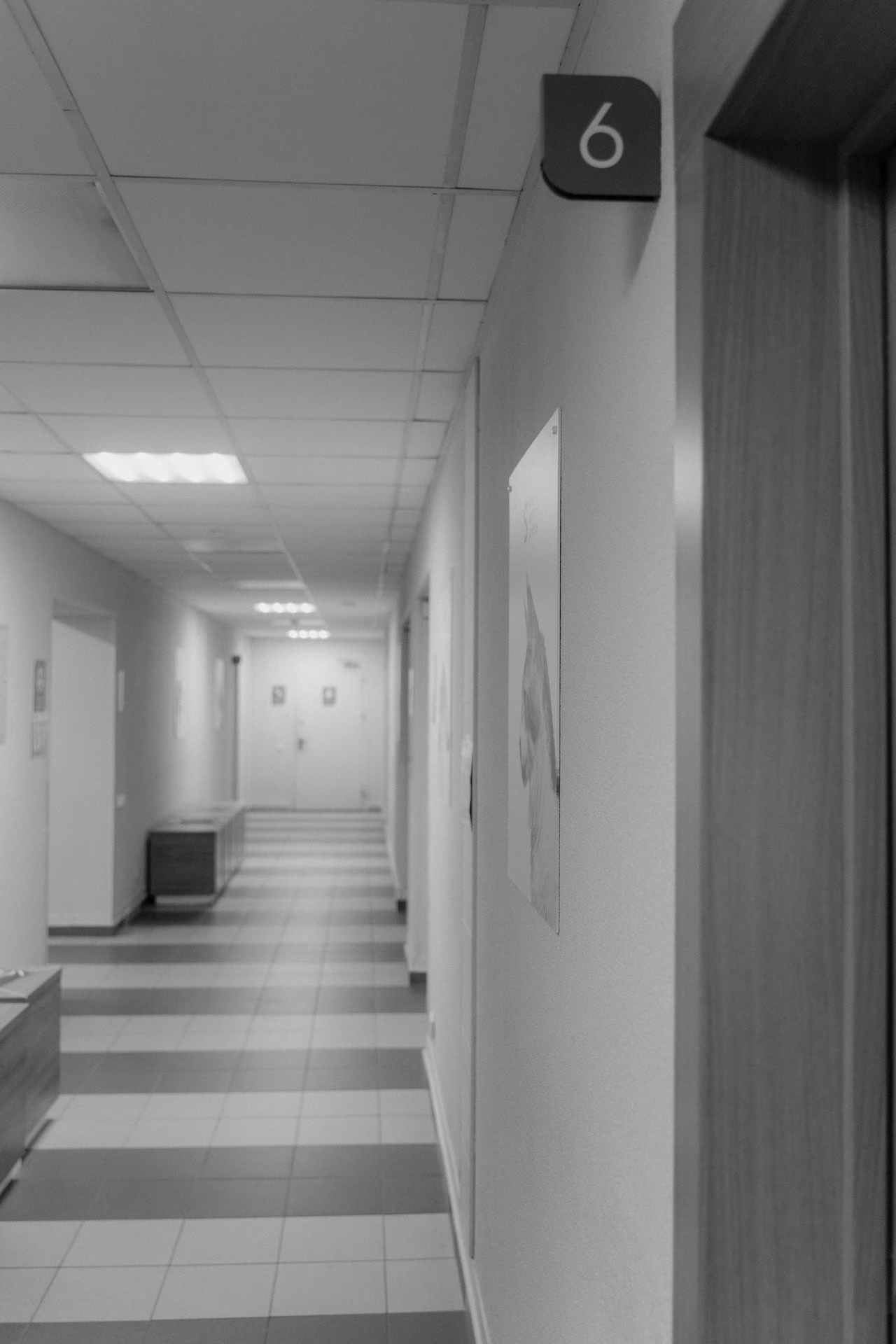 A grayscale corridor in a hospital showing an empty hallway with a room number sign.