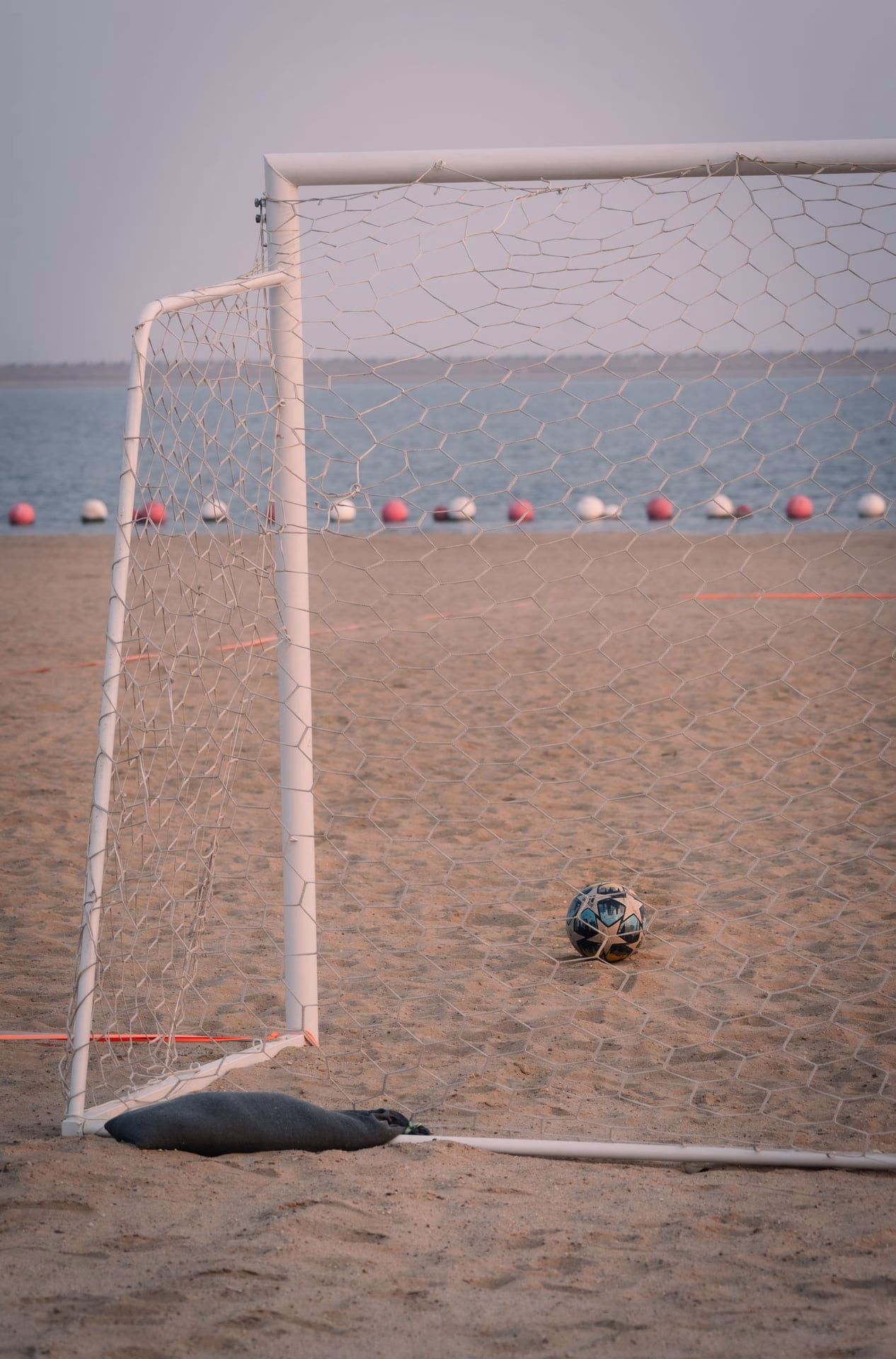 A soccer ball near a goal on a sandy beach by the sea at sunset, evoking a relaxed sporting atmosphere.