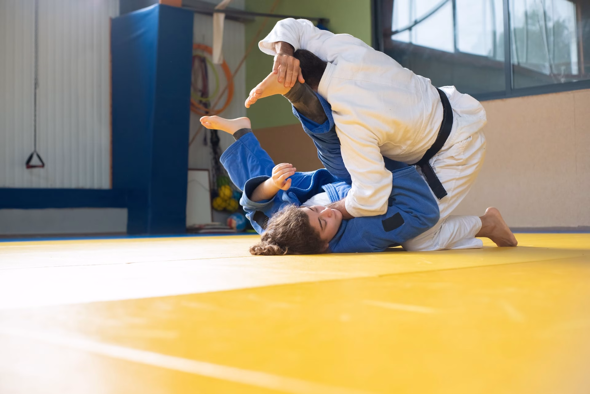 A man and woman engage in judo training on a yellow tatami mat indoors.