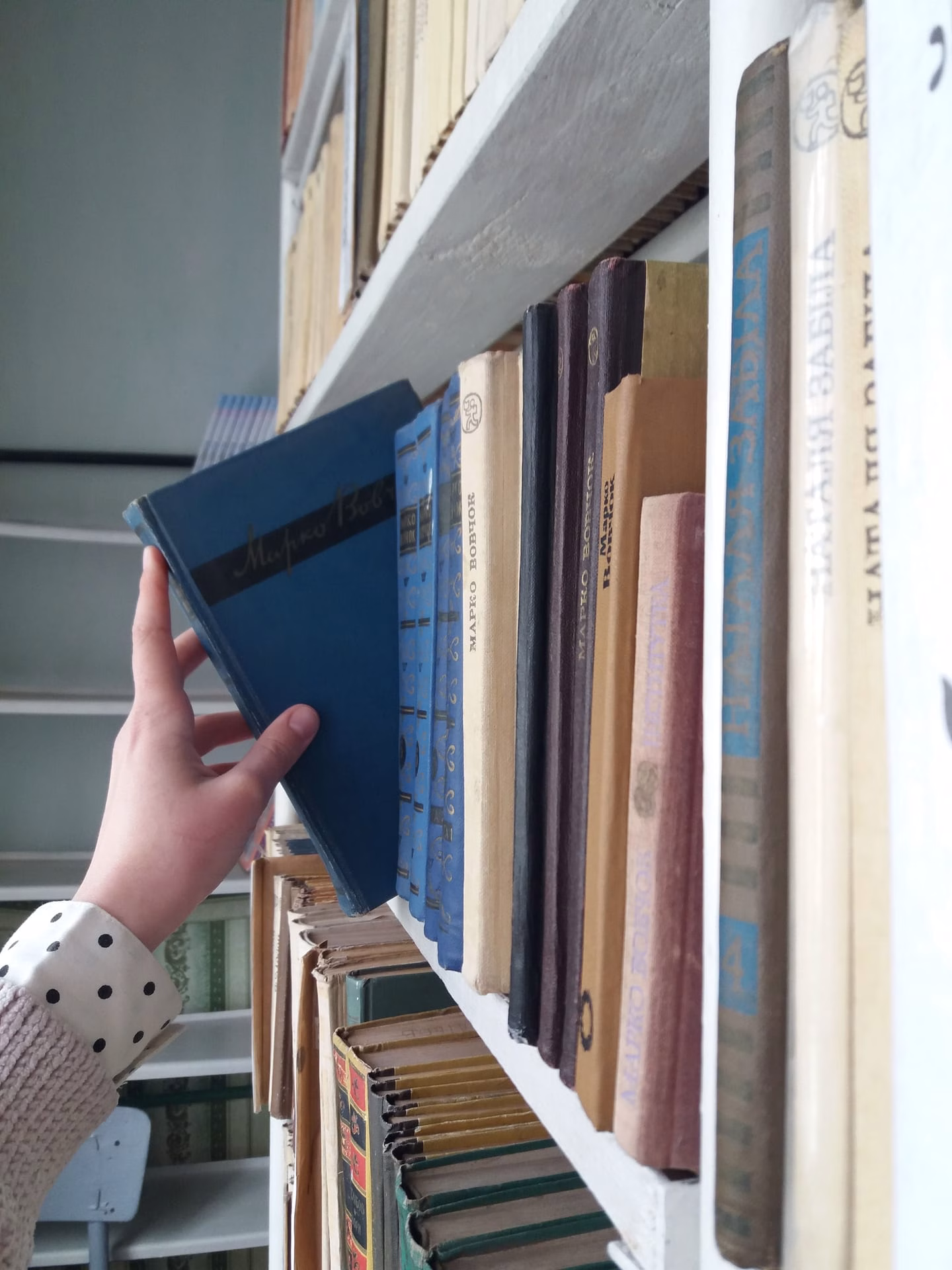 A hand reaching for a blue book on a library bookshelf with various literary titles.
